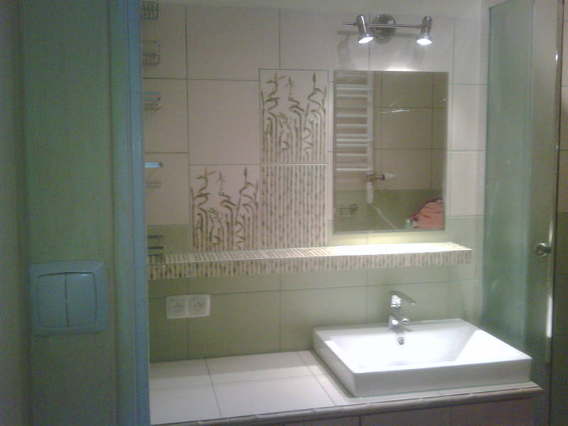 Wykończona łazienka z kwadratową umywalką, baterią oraz płytkami w odcieniach bieli i zieleni z motywem roślinnym, oświetlona dwoma punktowymi lampami nad lustrem.